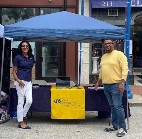 A female dressed in a dark polo shirt and a male wearing a light yellow sweater are standing in front of a blue canopy tent and table draped with a College of Nursing and Health Professions table cloth.